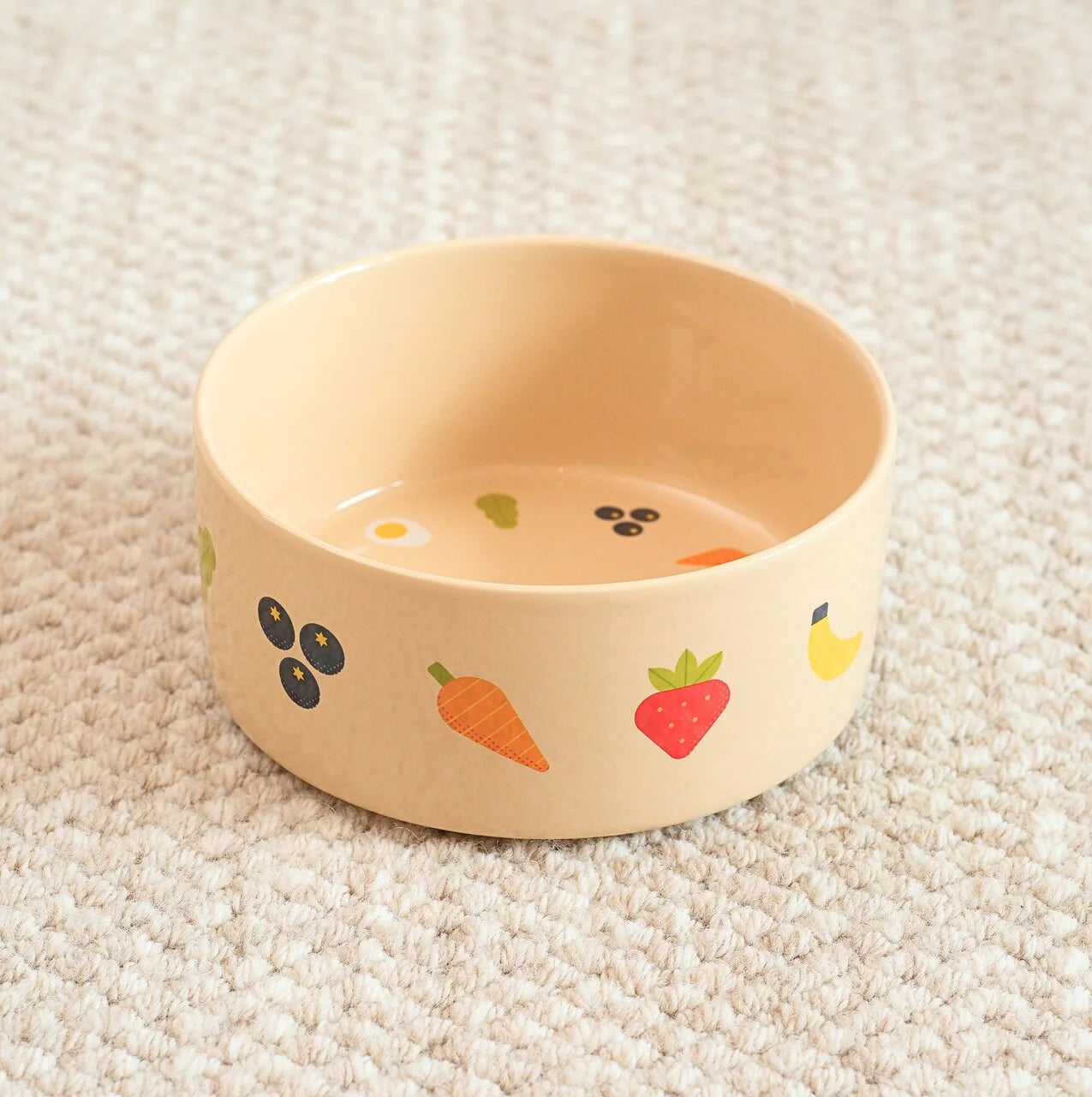 Beige ceramic dog bowl with fruit and vegetable patterns on a beige carpet